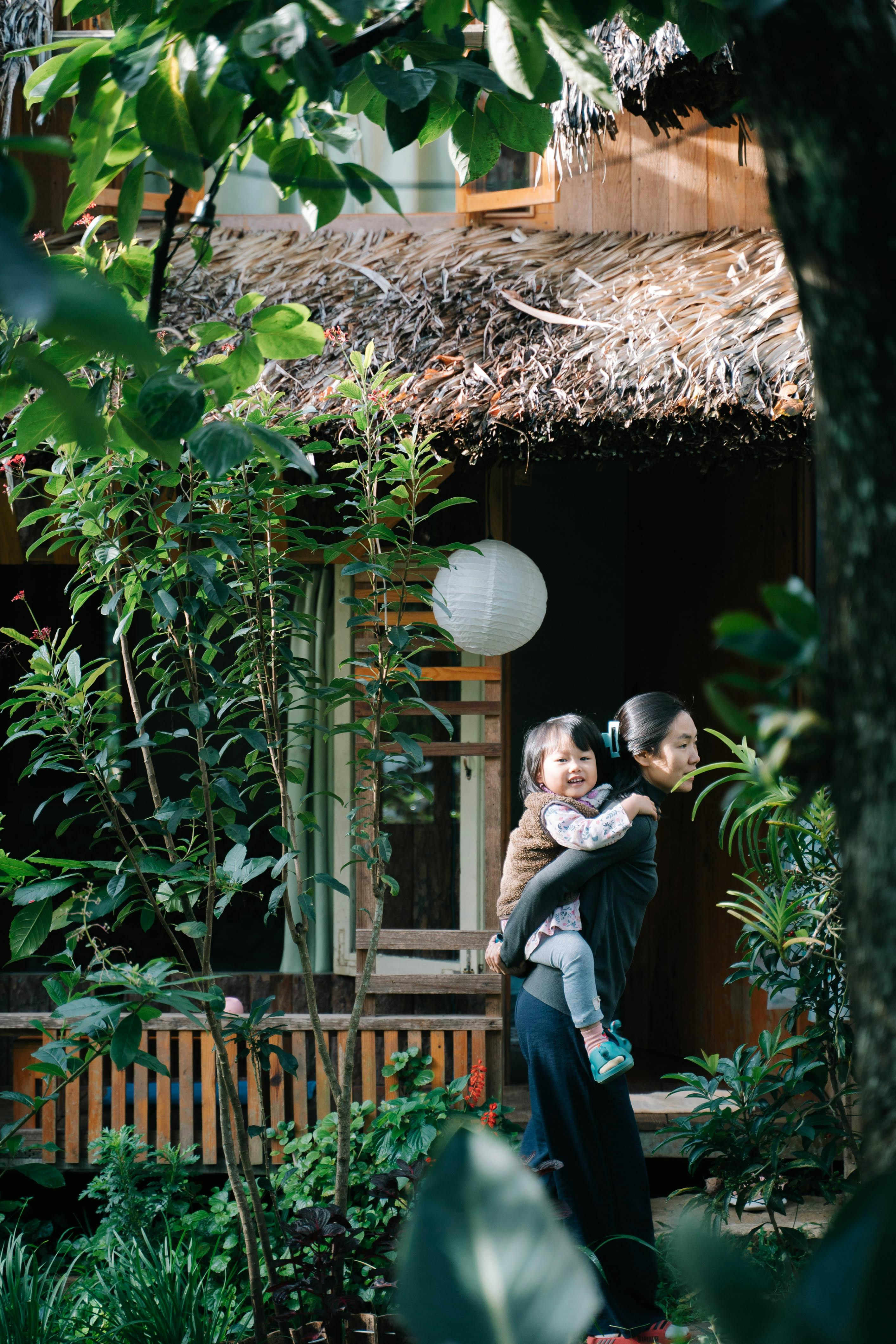 An Asian mother joyfully carries her child in a verdant garden by a thatched-roof house.