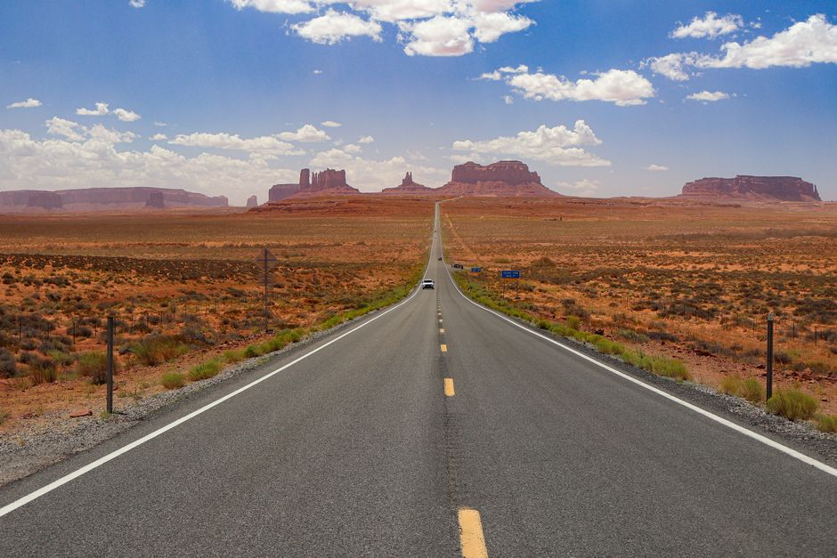 A long, empty highway stretches through Monument Valley under a bright blue sky.