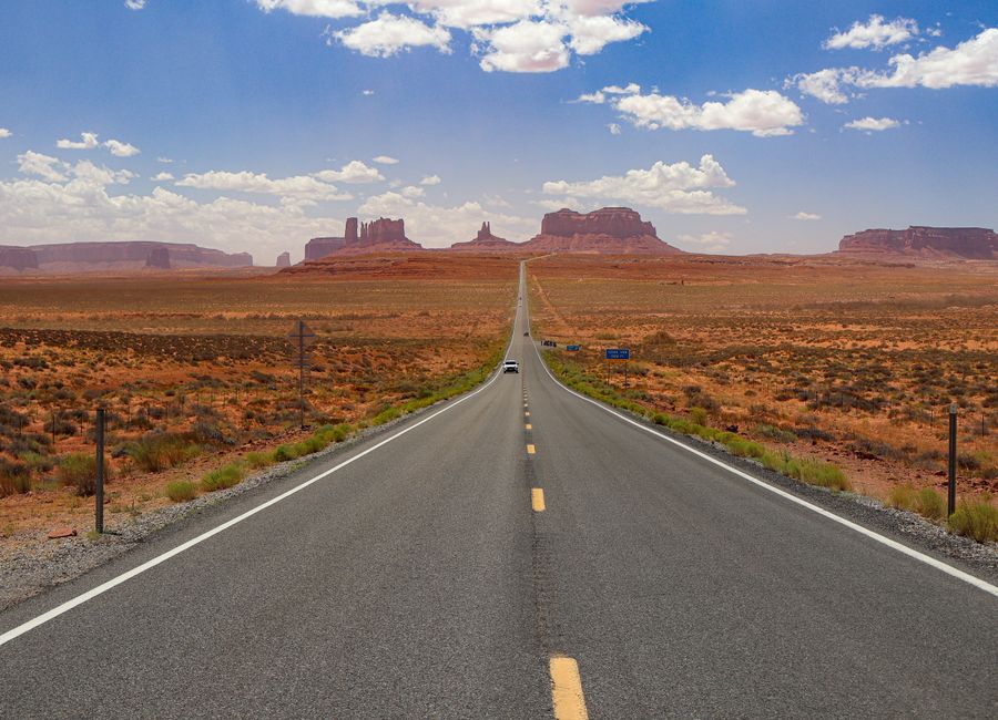 A long, empty highway stretches through Monument Valley under a bright blue sky.