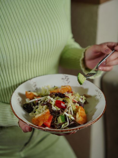 A woman in a green sweater holds a bowl of fresh vegetable salad with a fork indoors.