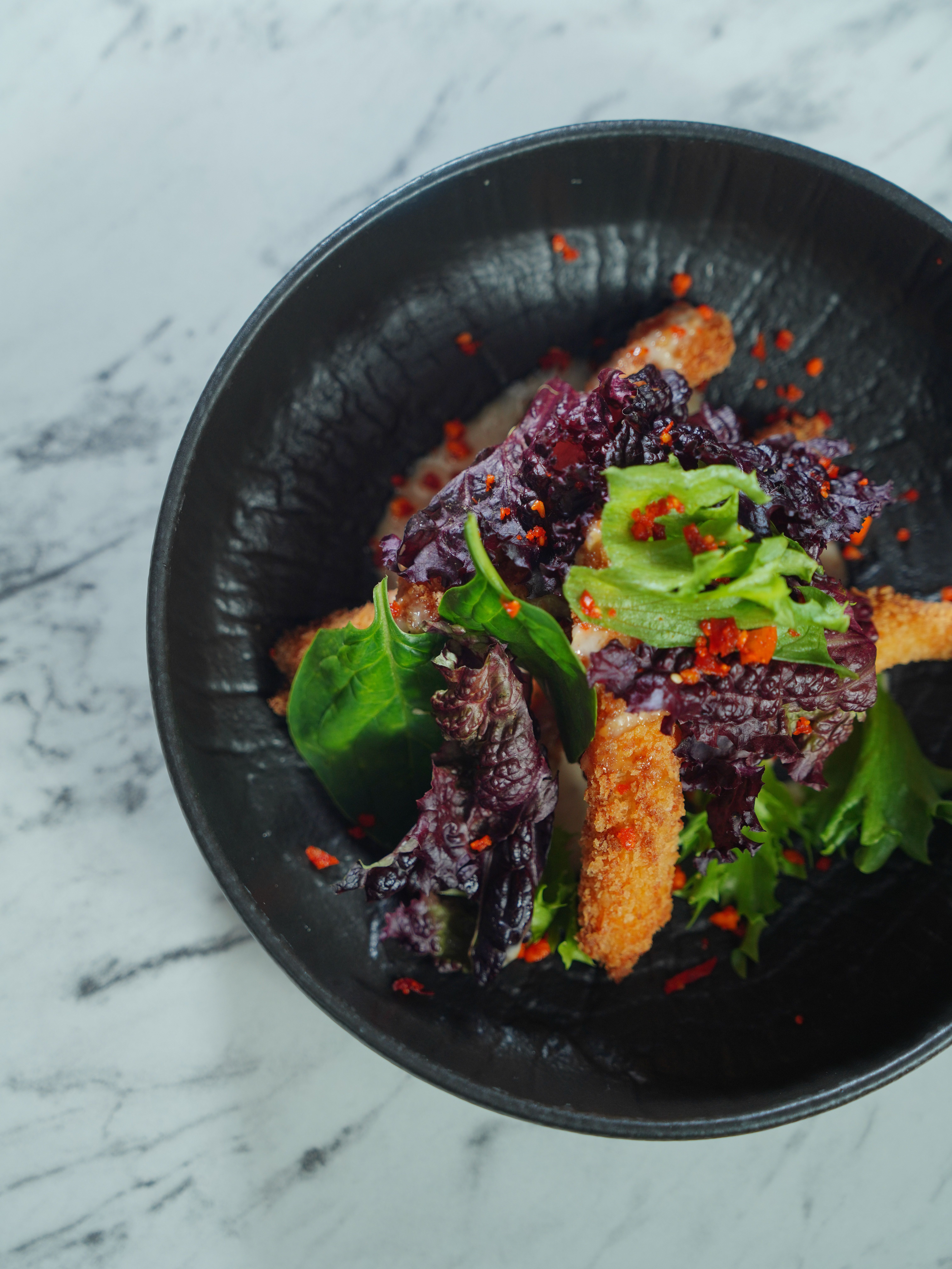 Top view of a crispy chicken salad with mixed greens in a stylish black bowl on marble surface.