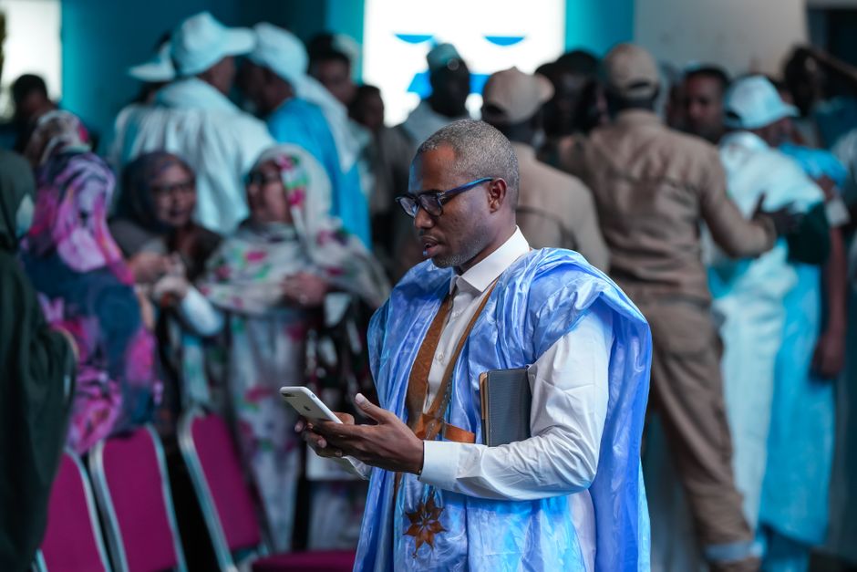 Man in traditional blue robe uses smartphone in crowded indoor event in Nouakchott, Mauritania.