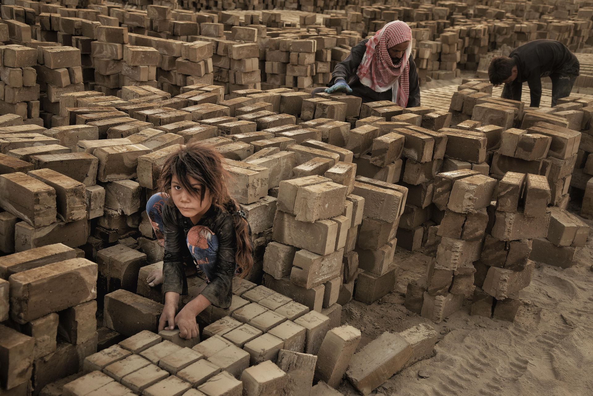 Child working in a brick factory surrounded by large stacks of bricks.