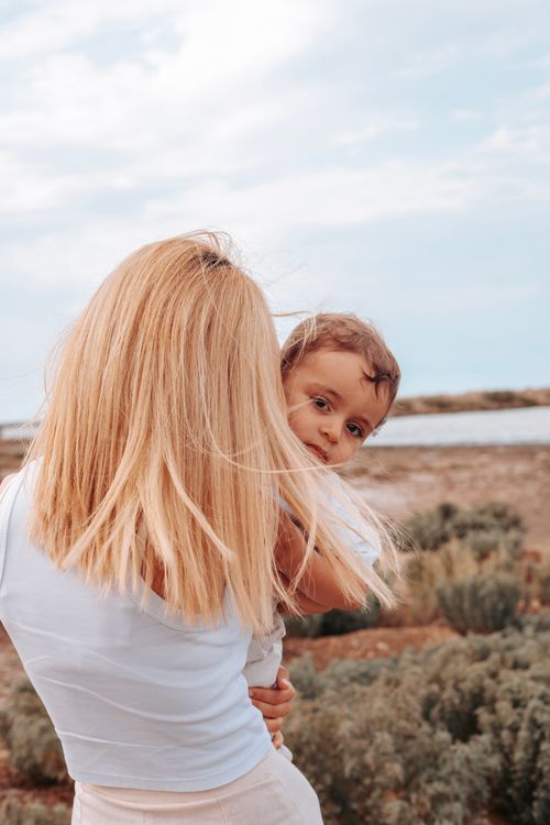 Free A mother with long blonde hair holds her child near the seaside, showcasing a warm family moment. Stock Photo
