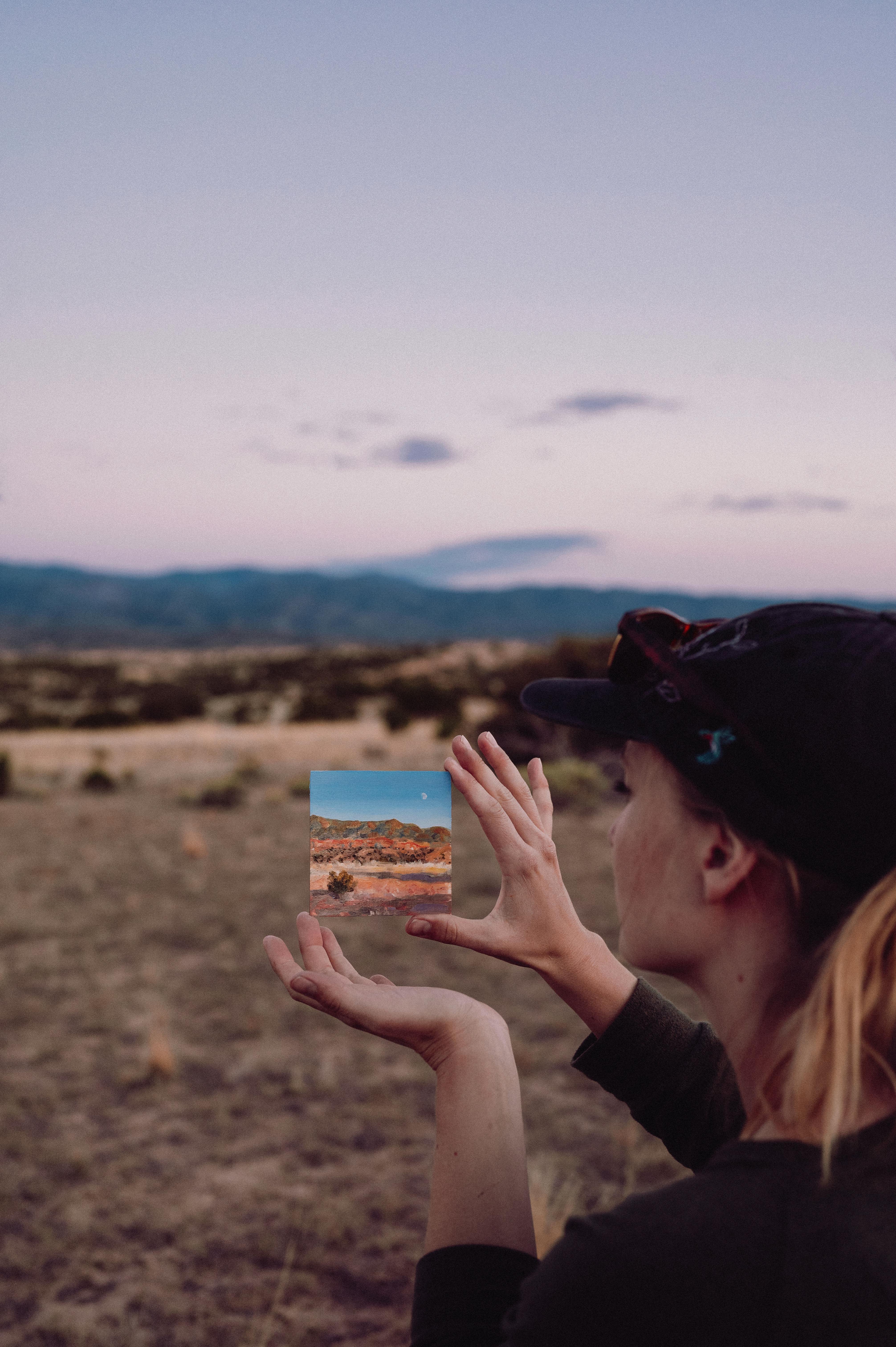 A woman holds a photo of a desert landscape against a scenic outdoor backdrop at dusk.