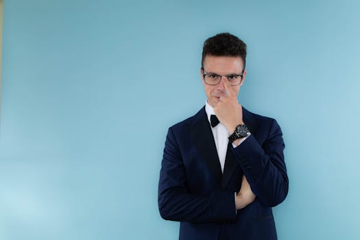 Elegant man in a navy blue suit with eyeglasses and wristwatch posing against a blue background.