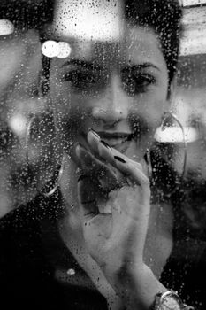 Black and white portrait of a smiling woman behind a rain-covered glass window.