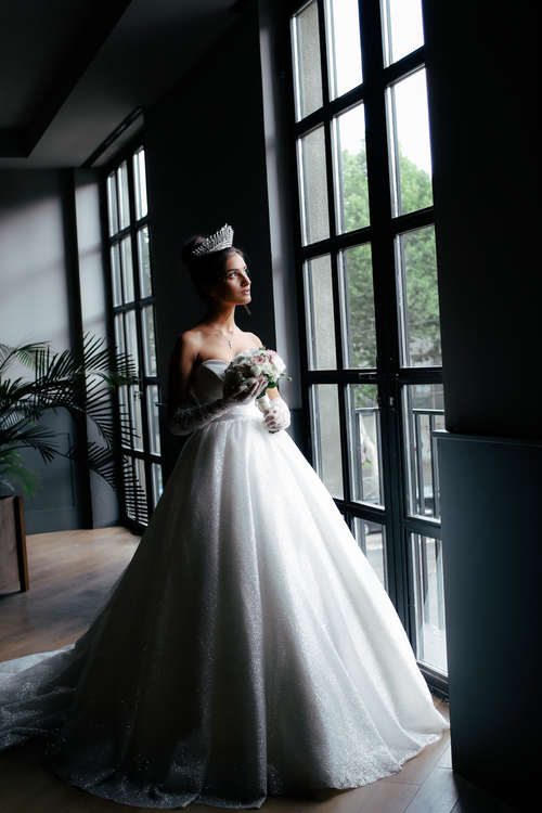 Free Bride in a sparkling strapless gown holding a bouquet by a large window, wearing a tiara. Stock Photo