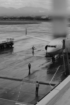 A dramatic black and white image of a runway with an airplane and workers on a rainy day.