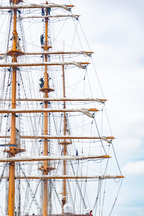 Free Sailors man the rigging of a classic tall sailing ship with wooden masts against a clear sky. Stock Photo