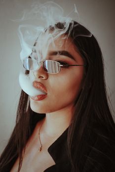 Portrait of a stylish woman exhaling smoke, wearing sunglasses indoors.