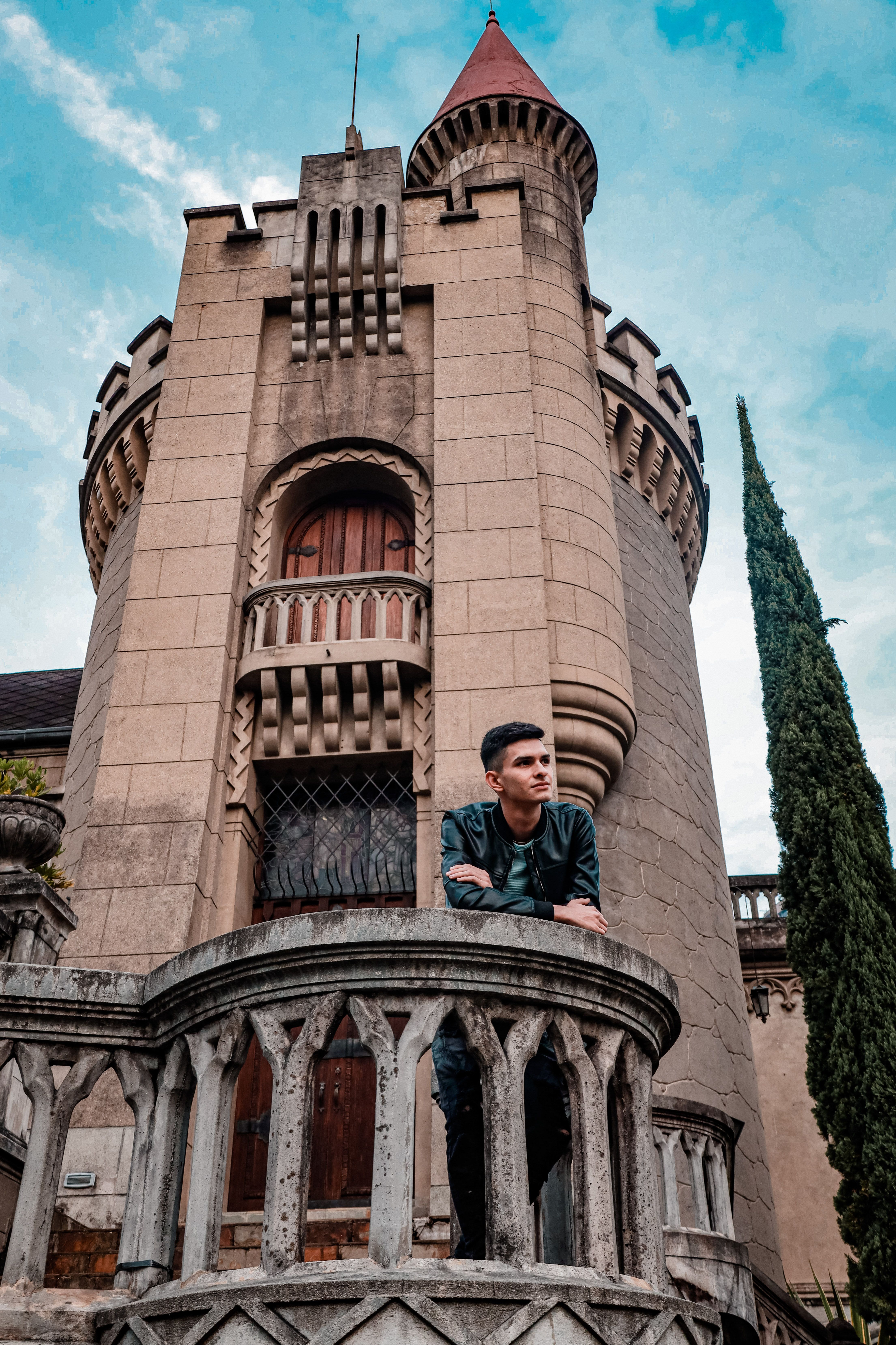 A young man leans on a historic castle balcony, showcasing its intricate architecture under a bright sky.