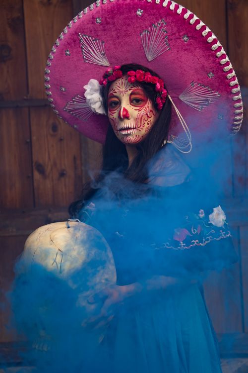 Free Woman in traditional Day of the Dead costume with face paint and pink sombrero holding a large skull amidst blue smoke. Stock Photo