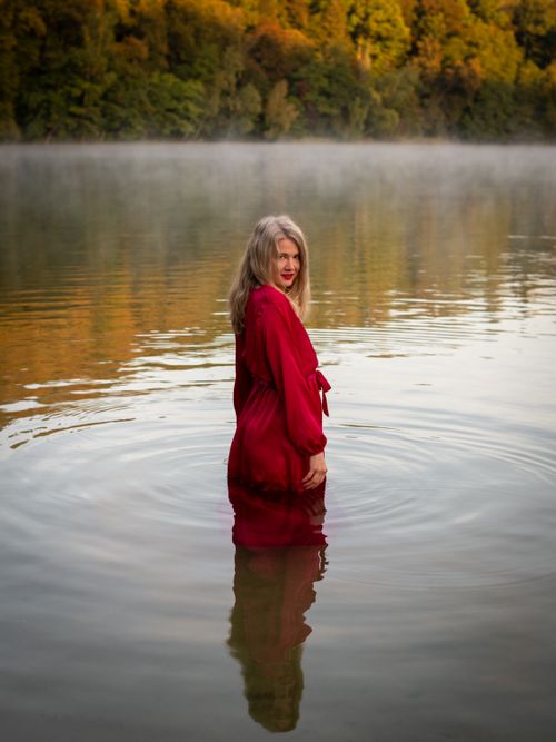 Free Woman in a red dress standing in a tranquil autumn lake in Lithuania. Stock Photo