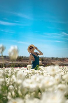 A smiling woman in a straw hat stands in a vibrant flower field under a clear blue sky.