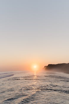 A peaceful sunrise over the ocean with misty cliffs in the background, captured in a vertical shot.