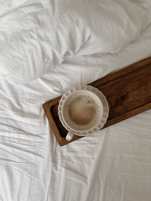 Top view of a coffee cup on a wooden tray on white bed sheets.