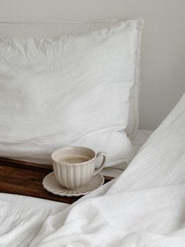 Warm cup of coffee resting on a wooden tray amidst white linens for a cozy morning vibe.