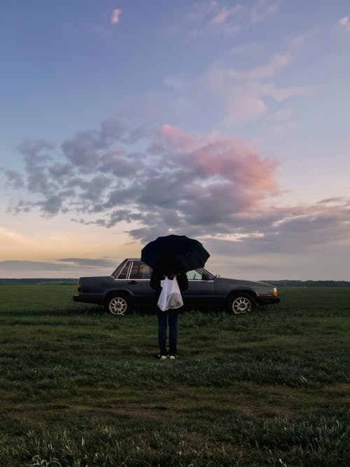 Person with Bag and Umbrella Standing near Vintage Car on Grassland