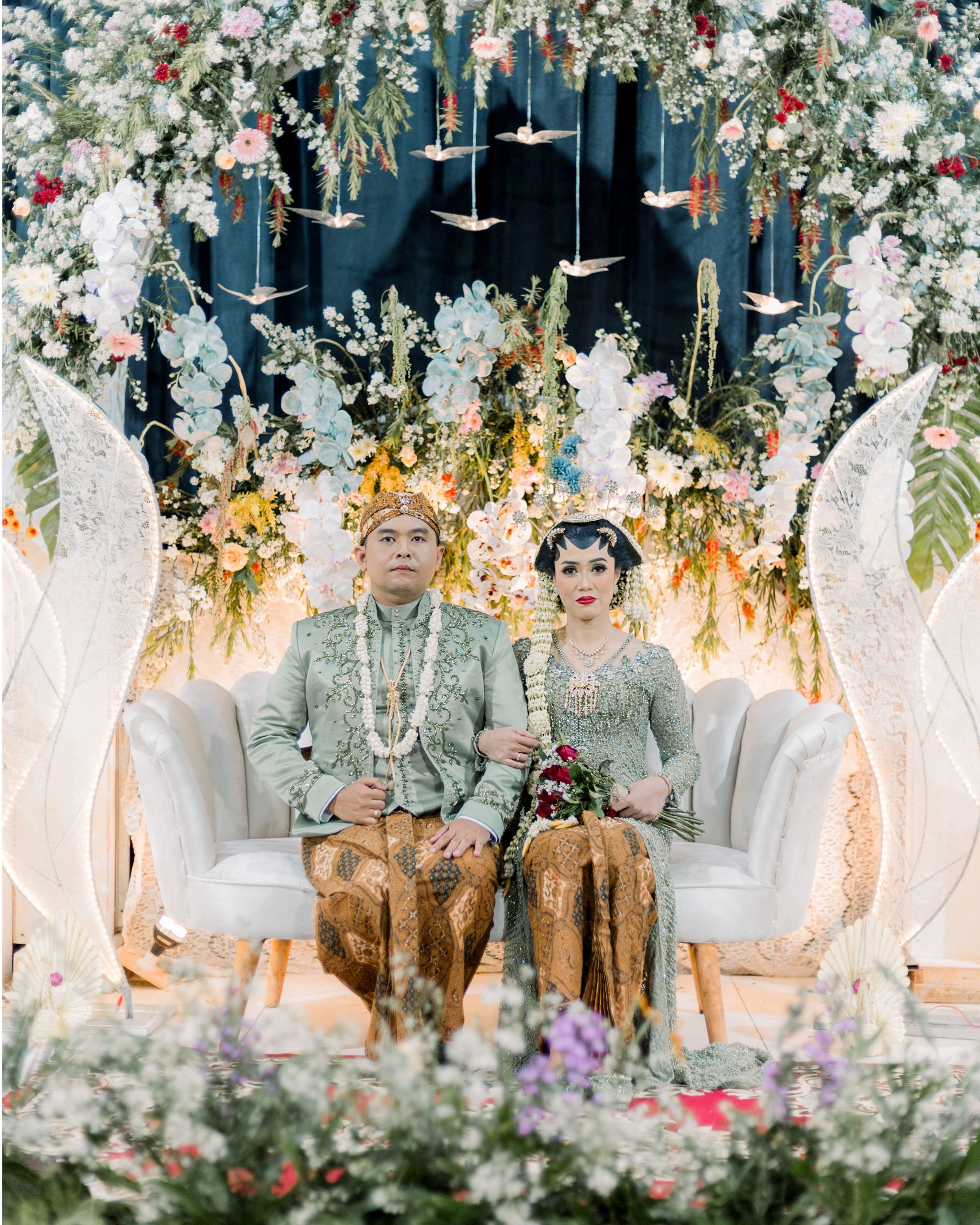 Traditional wedding couple seated in ornate floral setting in a cultural ceremony.