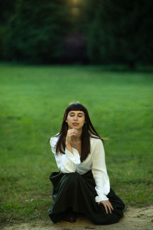 Free A young woman sits elegantly with eyes closed in a serene park, wearing a white shirt and dark skirt. Stock Photo