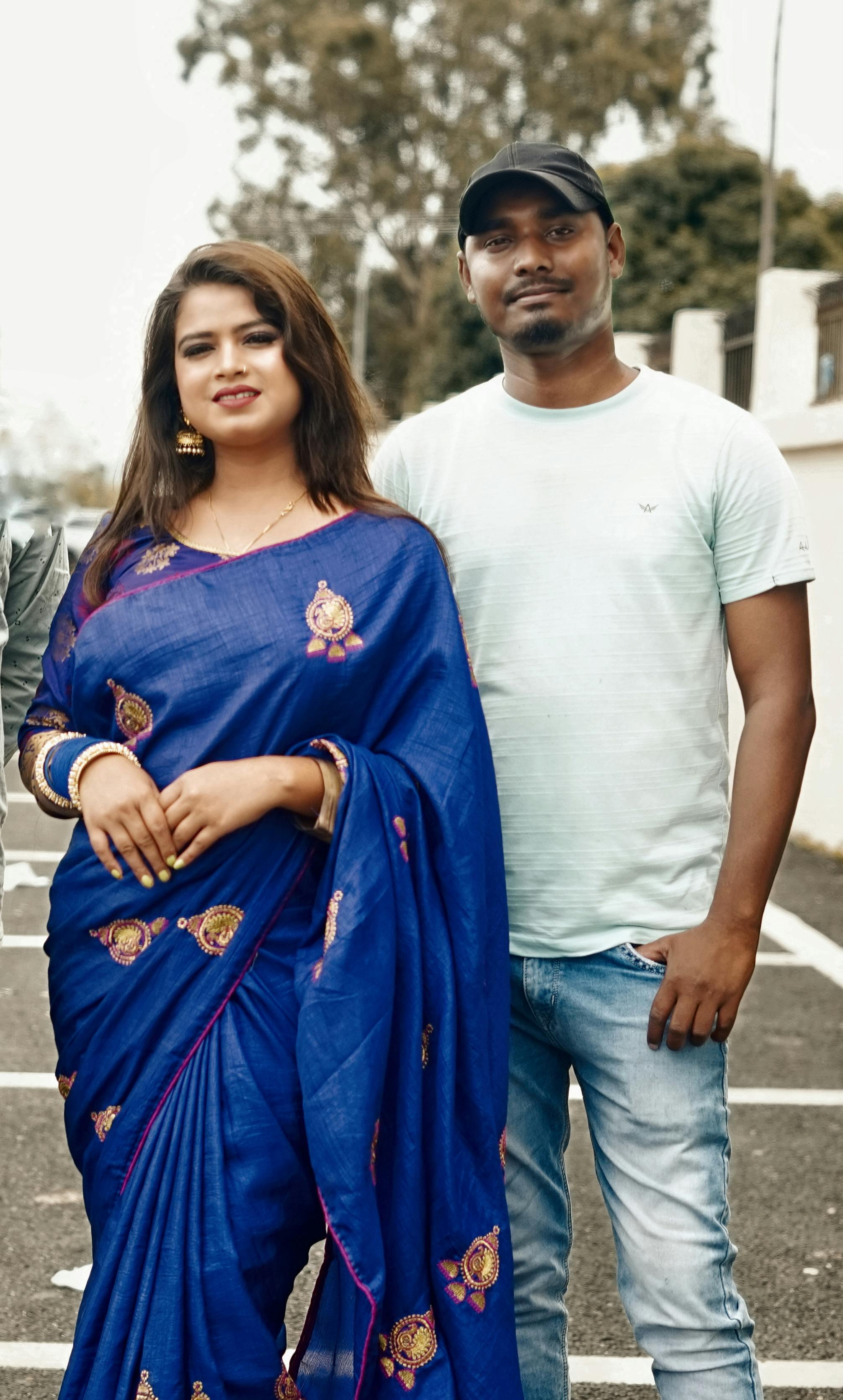 A couple stands outdoors, with the woman in a traditional blue sari and the man in casual attire, embodying a blend of cultures.