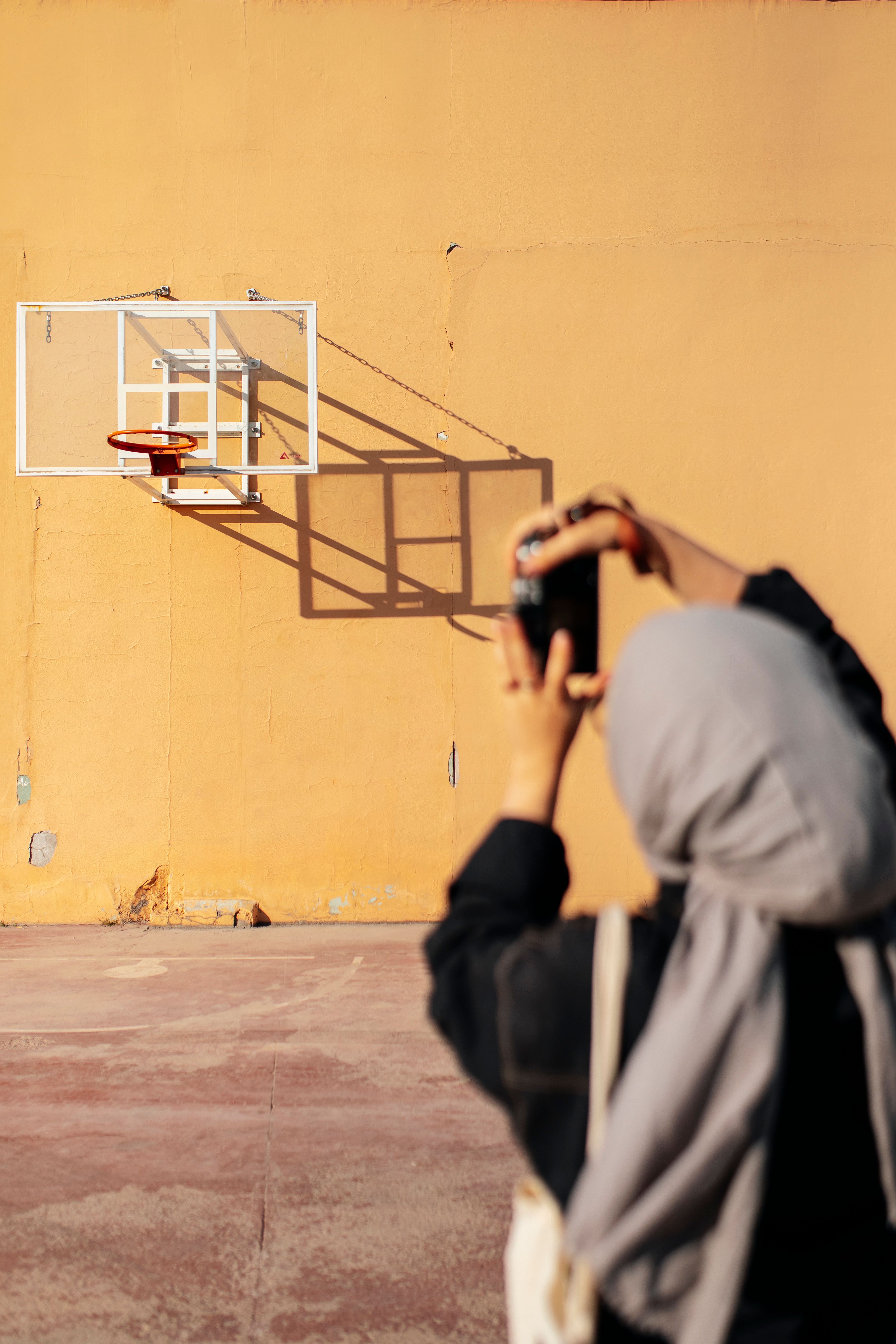 A woman captures a basketball hoop on a sunny day, highlighting urban sports culture.