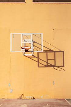 A basketball hoop casting a long shadow on a sunlit urban wall, embodying a minimalist aesthetic.
