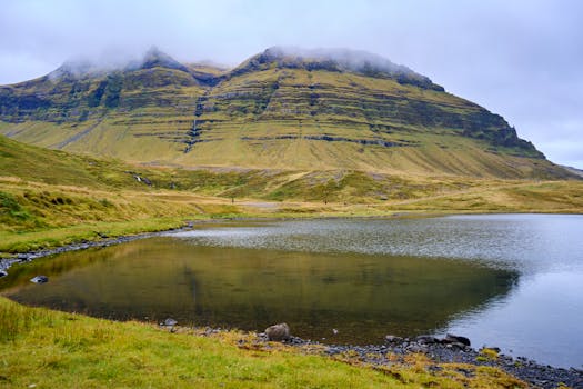 A serene landscape featuring a placid lake and green, eroded hills under a cloudy sky.