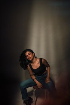 Moody studio portrait of a woman in a black top and jeans sitting on a stool.
