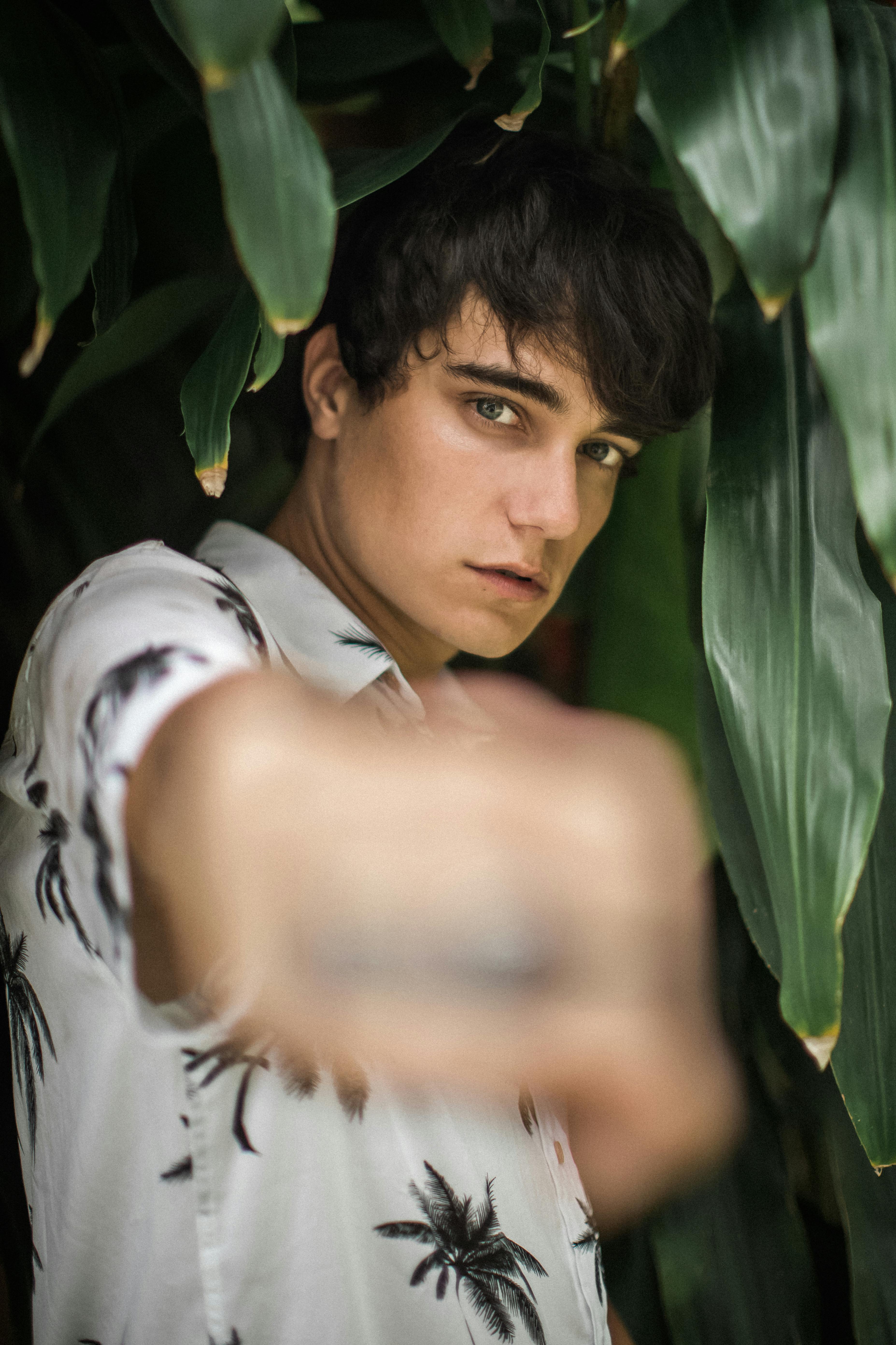 A contemplative young man in a tropical shirt stands amidst lush green foliage, striking a casual pose.