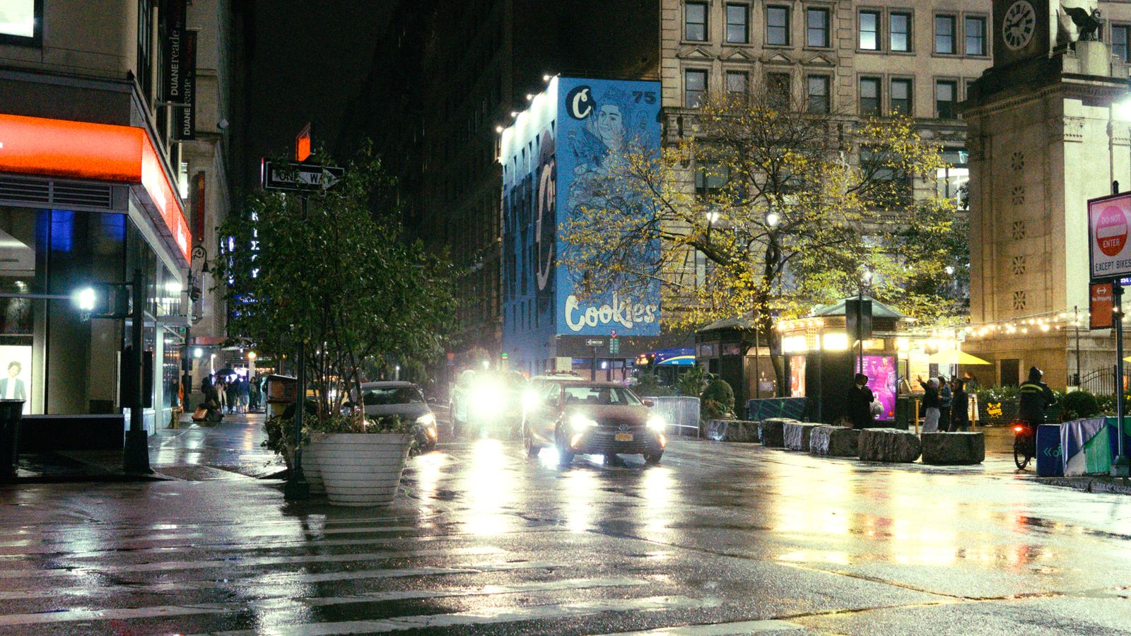 Nighttime city street view featuring bright headlights and illuminated storefronts in an urban setting.