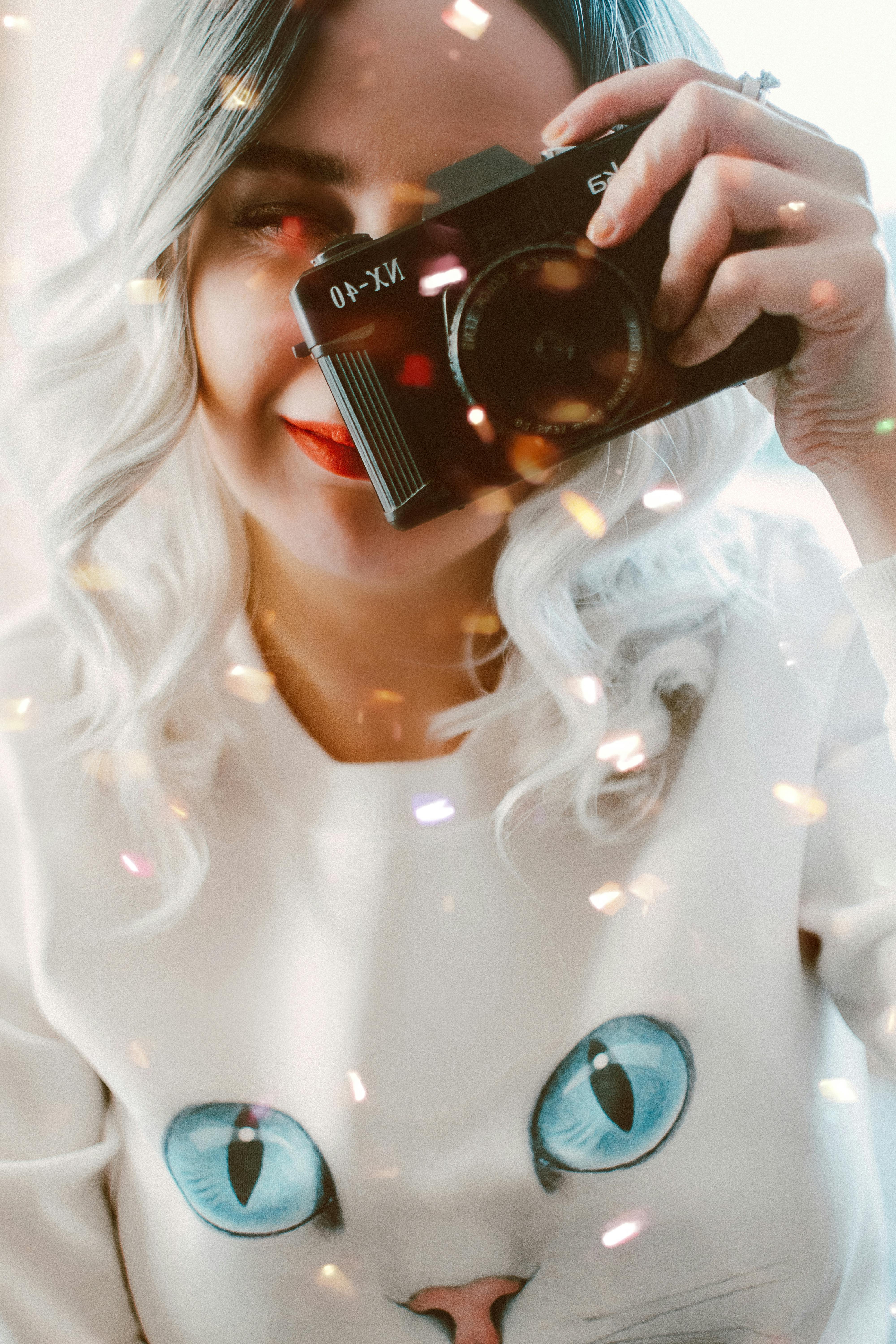 Woman capturing a photo with a vintage camera, wearing a cat print shirt, amid bokeh lights.