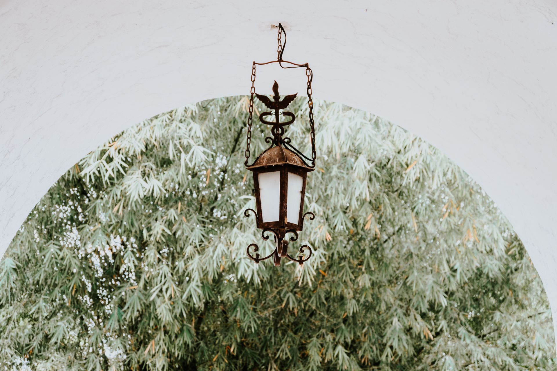 A rustic vintage lantern hanging beneath an arched alcove with lush greenery in the background.