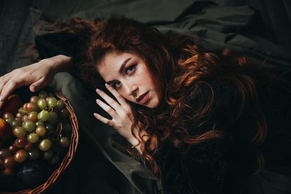 Moody indoor portrait of a woman with red hair lying next to a fruit basket, showcasing elegance and grace.