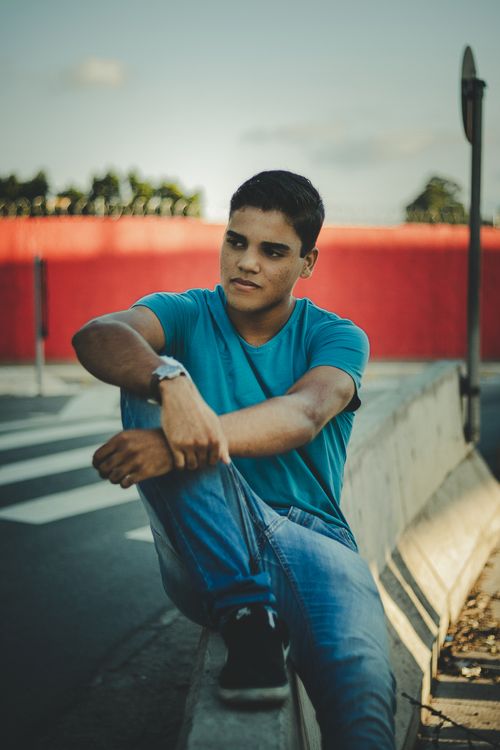 Free A young man in casual wear sits outdoors by a roadside barrier, exuding a relaxed vibe. Stock Photo