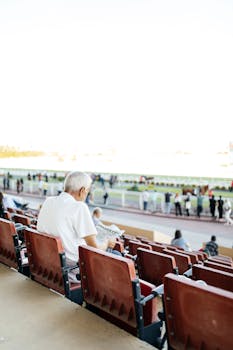 Senior man in stadium seating reads newspaper during outdoor sports event.