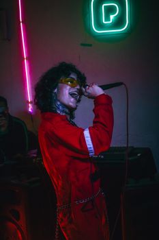 A lively singer in vibrant red attire delivers a powerful performance in a neon-lit room.