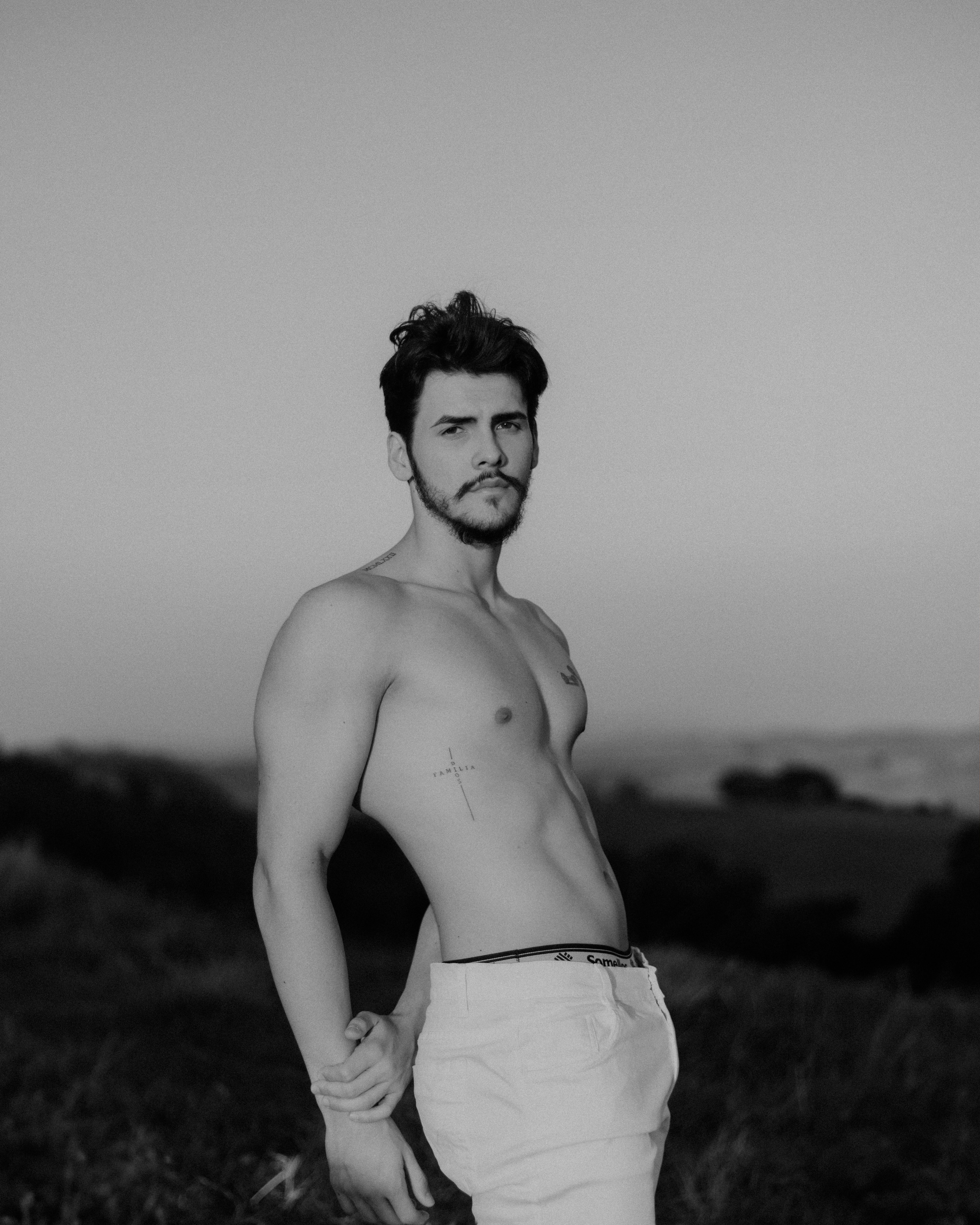 Dramatic black and white portrait of a topless man with tattoos in an outdoor setting.