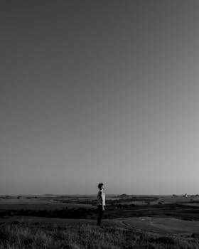 A lone individual stands in a vast rural landscape under a clear sky, evoking solitude.
