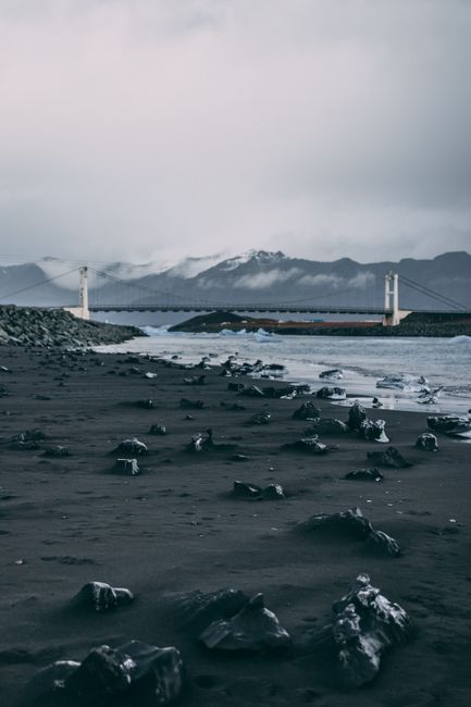 Stunning coastal view featuring a bridge, black sand beach, and distant snow-capped mountains.
