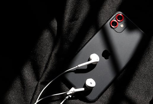 Black smartphone with white earphones on fabric, low-key lighting enhances modern technology vibe.