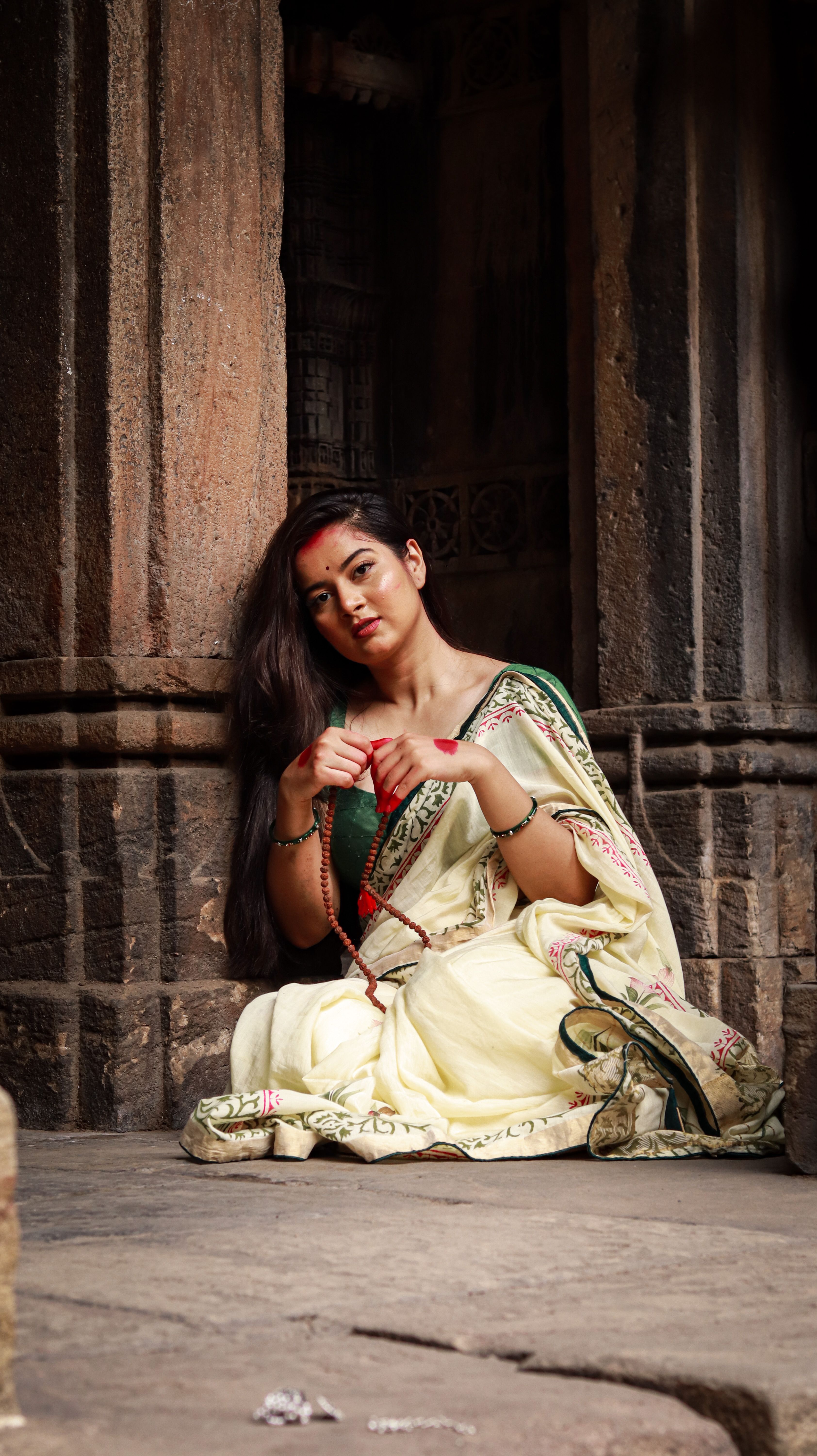 A young Indian woman in a saree sits gracefully against a weathered wall, embodying traditional elegance.