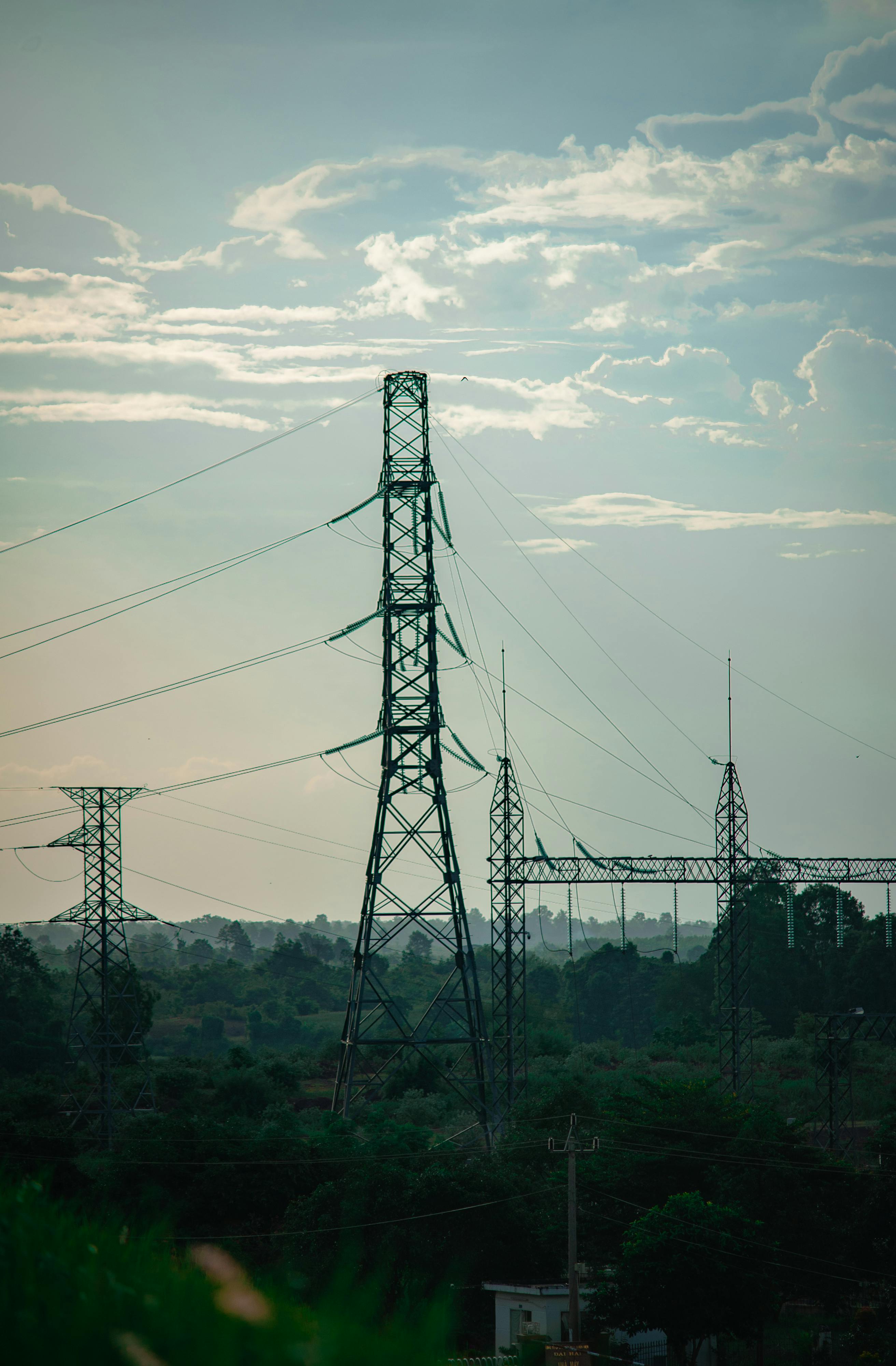 High-voltage power lines and towers in a rural setting under a cloudy sky.
