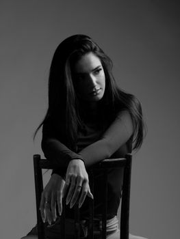 Artistic black and white portrait featuring a young woman sitting on a chair in a studio.