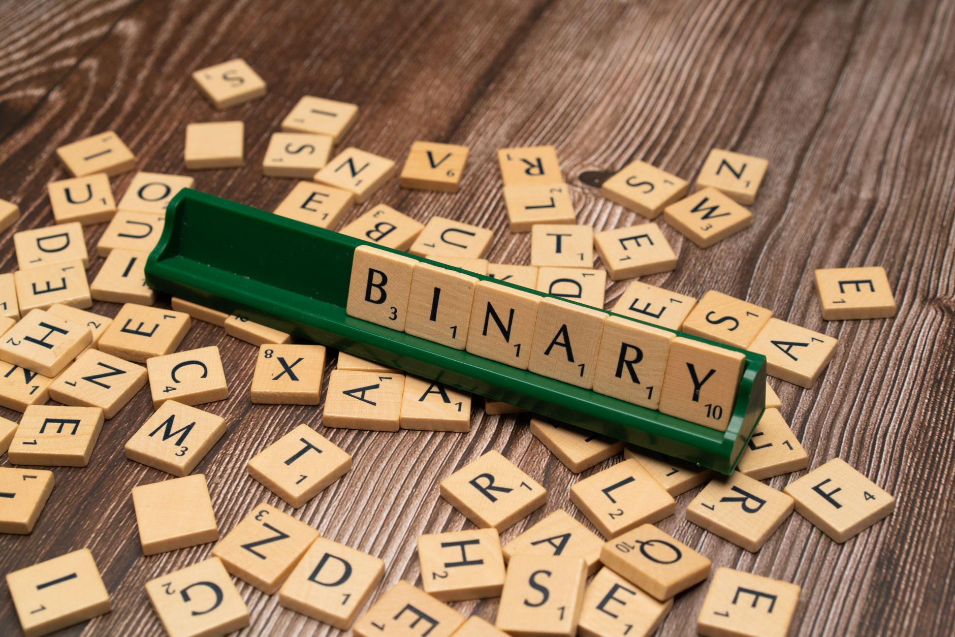 Close-up of scrabble tiles spelling 'Binary' surrounded by scattered letters on a wooden background.