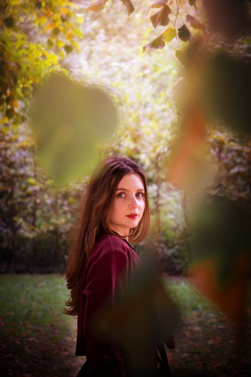Free Elegant portrait of a woman with long brown hair standing amidst lush greenery in a park. Stock Photo