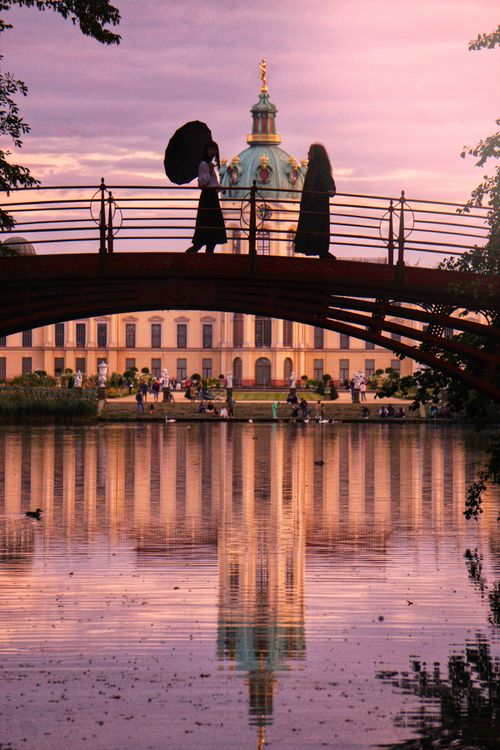 Free Silhouetted figures on footbridge at sunset, palace and reflection create serene scene. Stock Photo