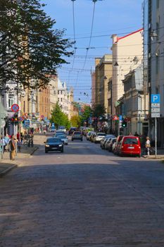 Urban scene of Riga, Latvia with cobblestone street, parked cars, and pedestrians on a sunny day.
