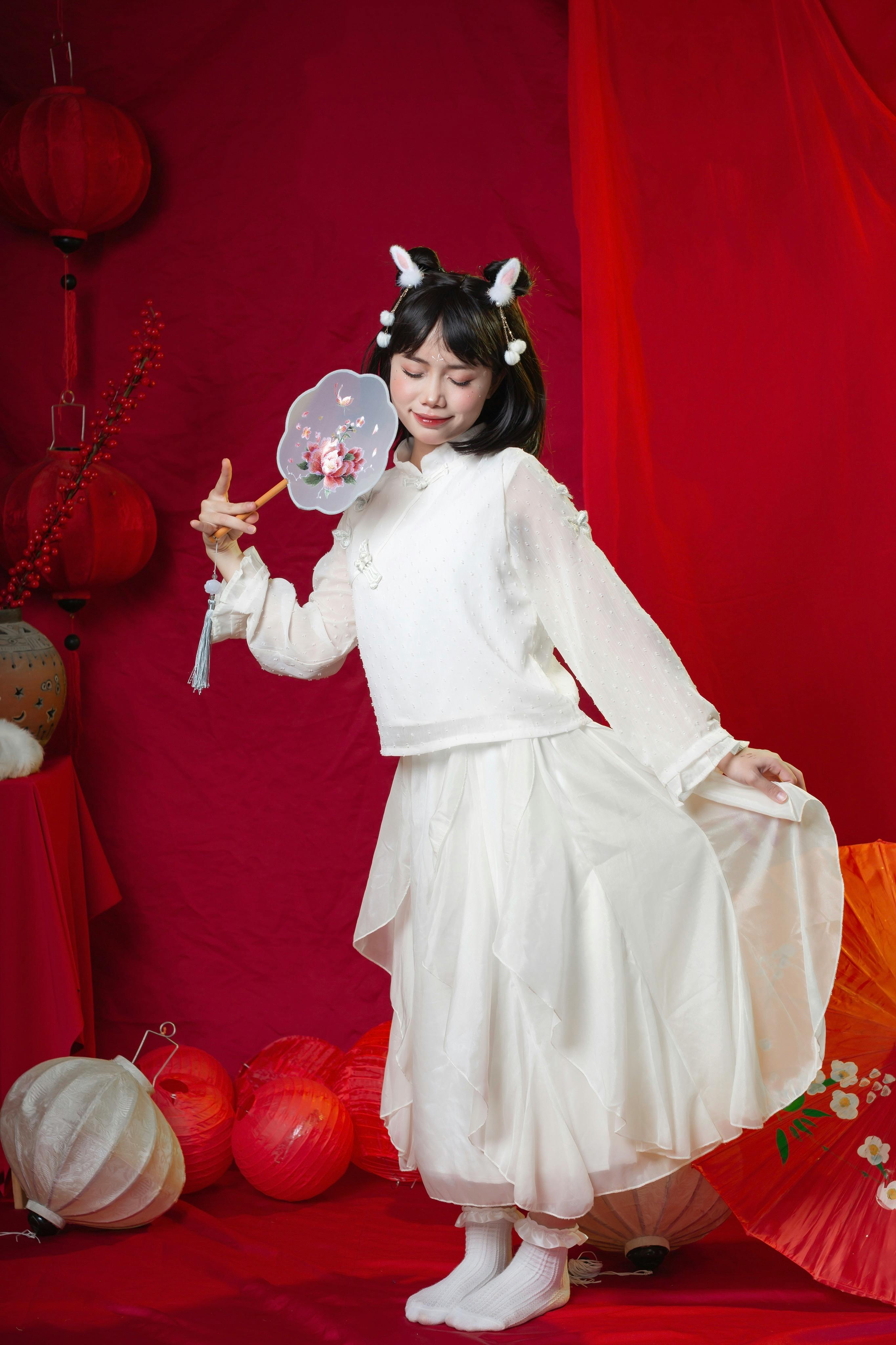 Asian woman in traditional white clothing posing with fan against a vibrant red background.